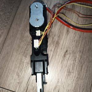 Electrical module with a small DC motor in a black plastic housing, connected to yellow and brown wire harnesses on a wooden floor description.