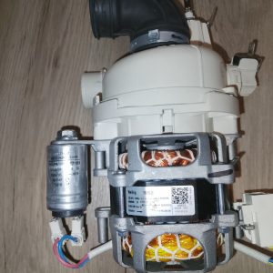 Motor pump assembly with white plastic housing and visible copper windings in a gray metal frame, mounted on a wooden floor. A black elbow pipe attaches to the top, with electrical wires and a small silver capacitor nearby. The label on the motor shows model information.