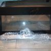 Brand-new stainless steel microwave oven with a black glass door, partially wrapped in plastic on a wooden floor.