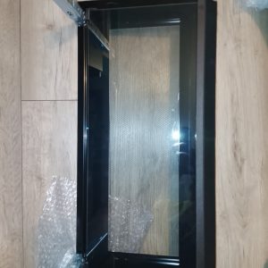 Black glass oven door with metal frame resting on a wooden floor, partially unpacked with bubble wrap seen nearby.