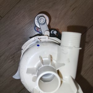 Close-up of a white plastic plumbing fitting with a metal mounting arm and bolt, on a wooden floor surface