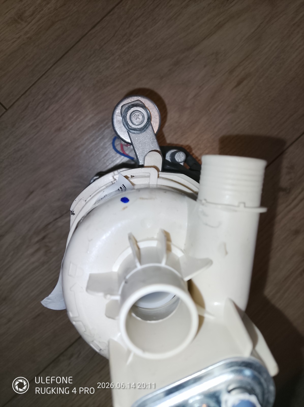 s-l1600-1159.jpg Close-up of a white plastic plumbing fitting with a metal mounting arm and bolt, on a wooden floor surface