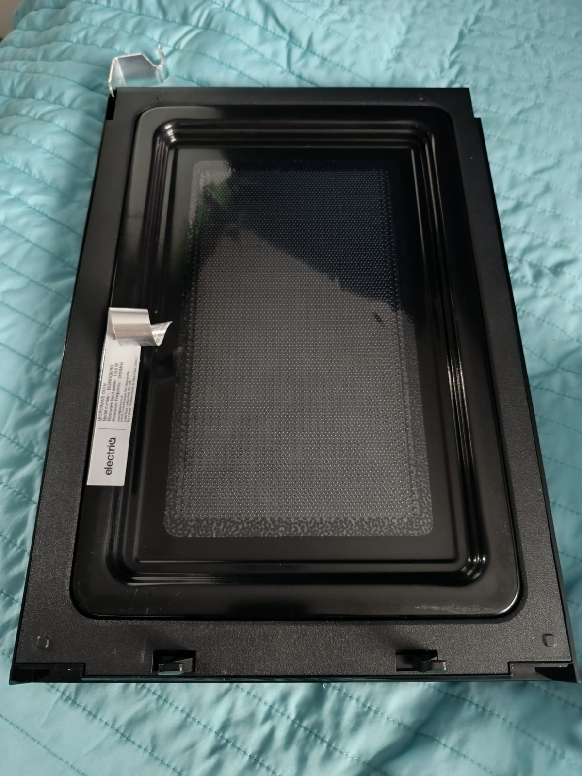 s-l1600-1233.jpg Black metal frame of a microwave oven door with an inner glass panel, placed on a blue quilted surface
