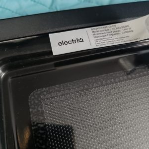 Close-up of a microwave oven interior with a white label showing model and power information attached to the door frame near a blue fabric surface beneath it.