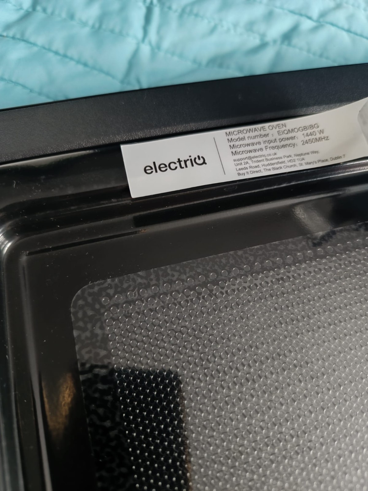 s-l1600-1234.jpg Close-up of a microwave oven interior with a white label showing model and power information attached to the door frame near a blue fabric surface beneath it.