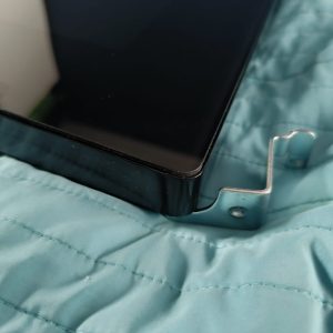 Close-up of a black tablet edge with a silver L-shaped mounting bracket attached to its corner on a blue quilted fabric surface.