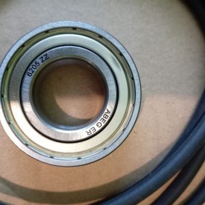 Close-up of a 6205 ZZ shielded ball bearing with inner and outer races, resting on a beige surface with a dark blue cable nearby.