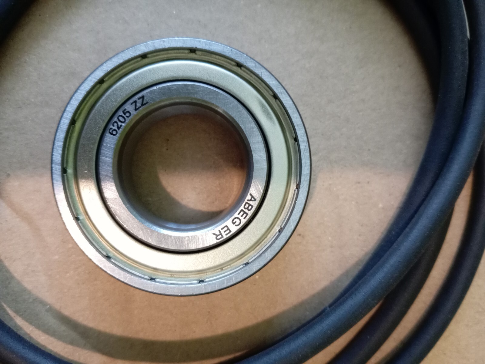 s-l1600-169.jpg Close-up of a 6205 ZZ shielded ball bearing with inner and outer races, resting on a beige surface with a dark blue cable nearby.