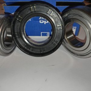 Three metal bearings resting on a white surface, with a blue box in the background. The center bearing has a black plastic inner element with grooves, flanked by two silver outer rings.