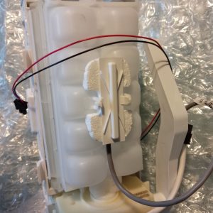 White plastic appliance component with red and black wires, mounted on a frame with foam padding and a silver foil backdrop.
