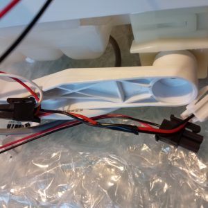 White plastic appliance component with a circular bore, connected to a red/black/white wiring harness and two connectors on a plastic bag surface.