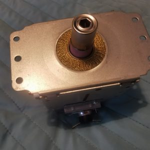 Close-up of a metal gearbox component: rectangular housing, central shaft, brass-textured seal ring, purple gasket behind it.