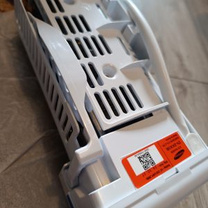 White plastic appliance part with vented grille, curved housing, and an orange label with a QR code on the surface (informational image)