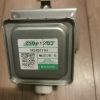 Metal electrical transformer with a green-labeled sticker reading M24B111H and a caution about high voltage microwave radiation, on a wooden floor