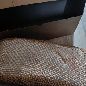 Open cardboard shipping box on a wooden floor, with a dark rectangular object inside and a honeycomb-patterned brown item in the foreground.