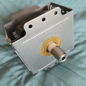 Close-up of a small metal mechanical assembly with a hollow shaft and a gold washer, showing a stack of leaf springs and vent holes in the casing.