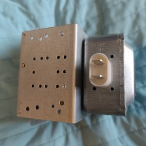 Close-up of a metal transformer with a perforated plate and a white two-prong plug outlet on the side.