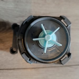 Close-up of a gray circular plastic component with a light blue four-spoke impeller in the center on a wooden surface.