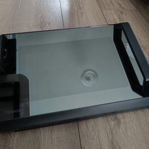 Toshiba flatbed scanner with glass lid lying on a wooden floor, reflection visible on glass surface.